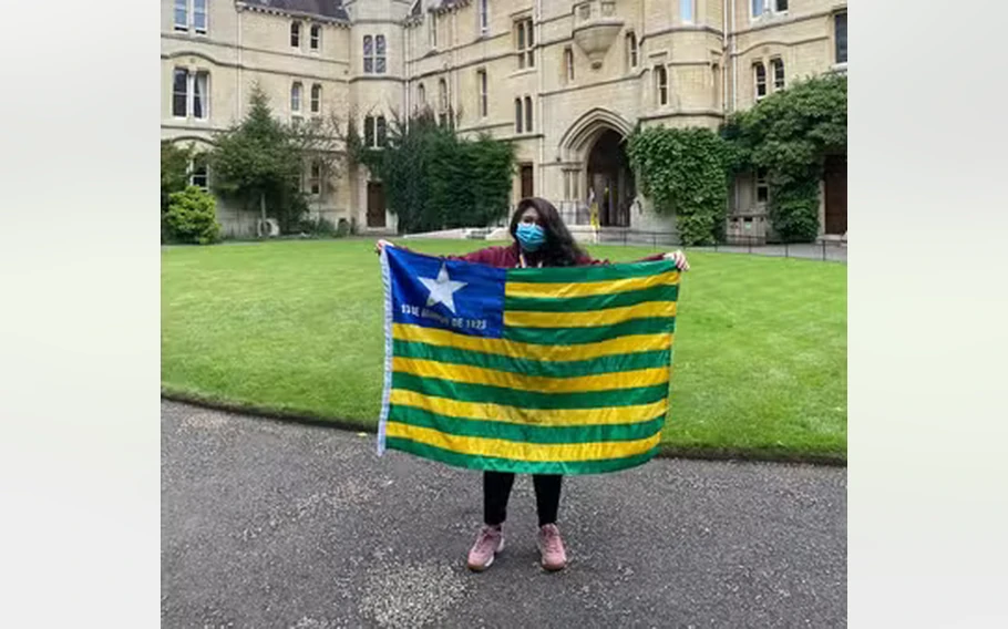Joanna Darck, de máscara azul, segura a bandeira do Piauí em frente a um edifício histórico, com gramado ao fundo.