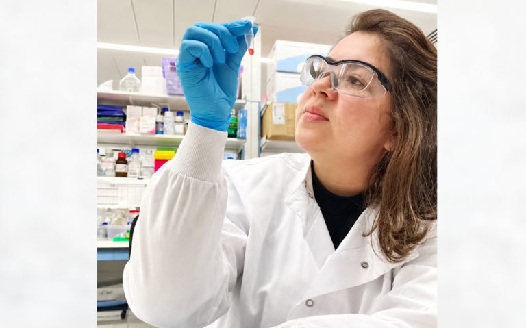 Joanna Darck em laboratório, usando jaleco, luva azul e óculos de proteção, enquanto observa um tubo de ensaio com líquido vermelho.