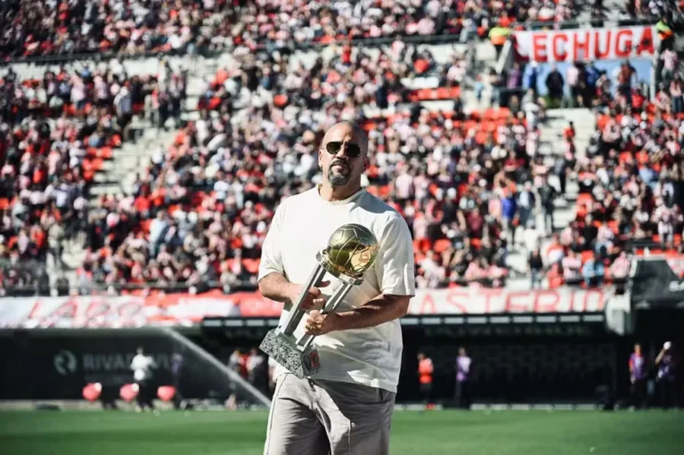 Juan Sebastián Verón, de camiseta branca e óculos escuros, segura um troféu dourado no gramado de um estádio, com torcedores nas arquibancadas ao fundo.