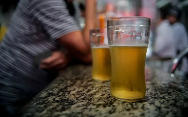 Dois copos com cerveja sobre um balcão de pedra, com pessoas desfocadas ao fundo.