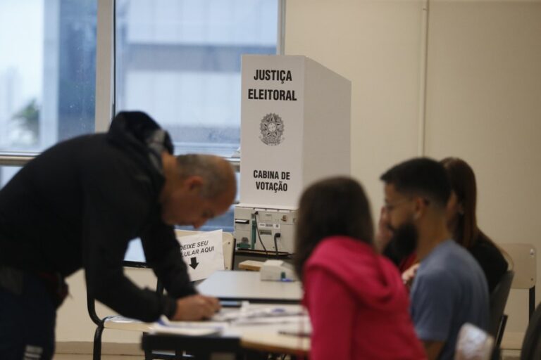 Homem inclinado sobre uma mesa em local de votação; ao fundo, urna eletrônica em cabine branca da Justiça Eleitoral e outras pessoas sentadas.