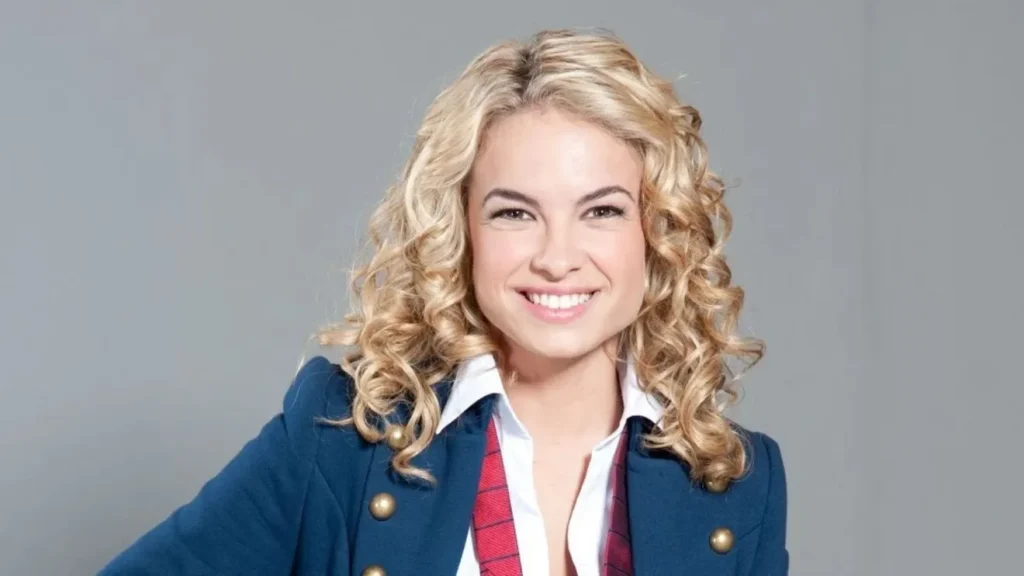 Lua Blanco com cabelos loiros cacheados, sorrindo, vestida com blazer azul, camisa branca e gravata vermelha, diante de fundo cinza.