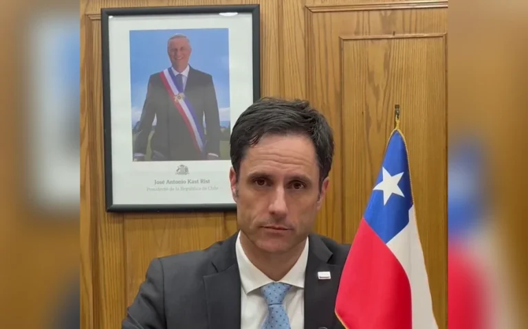 Luis Silva, de terno escuro e gravata azul, diante de uma bandeira do Chile e de um retrato presidencial em uma parede de madeira.