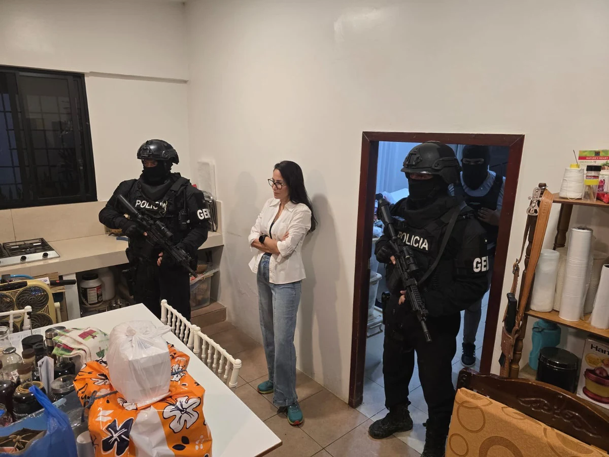 Luisa González de braços cruzados em uma residência, com policiais armados próximos.