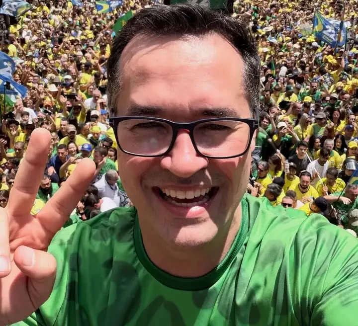 Homem de óculos e camisa verde sorri para uma selfie diante de multidão com bandeiras brasileiras.