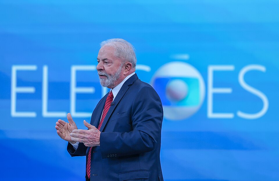 Lula, de terno azul e gravata vermelha, aparece de perfil, gesticulando com as mãos diante de um painel azul com o logotipo da TV Globo e a palavra Eleições.