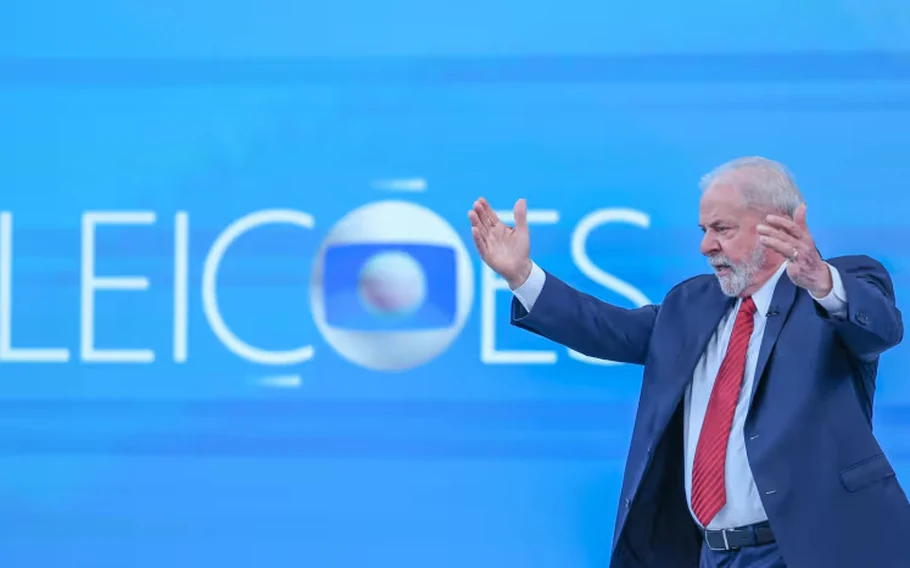 Lula, de terno azul e gravata vermelha, com os braços erguidos diante de um painel azul com o símbolo da TV Globo.