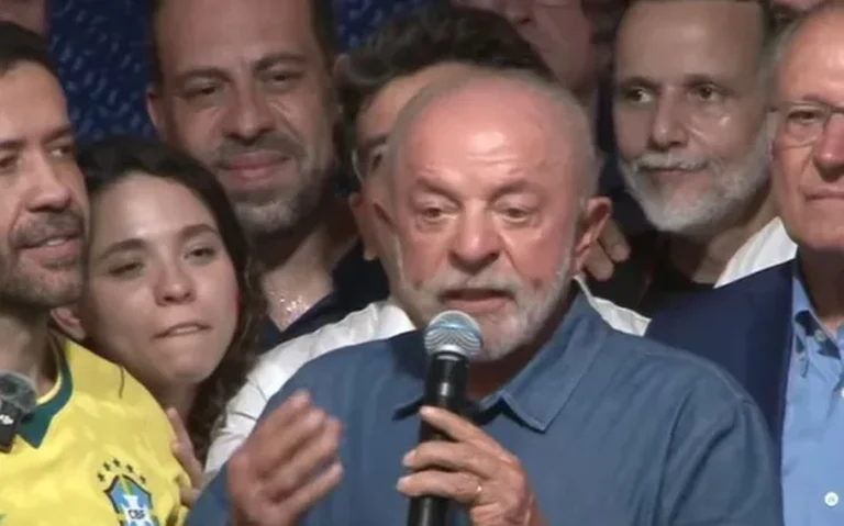 Lula, de camisa azul, fala ao microfone cercado por pessoas durante um discurso.