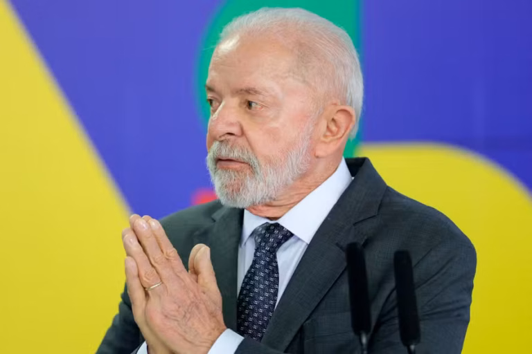 Presidente Lula de terno escuro e gravata, com as mãos juntas, diante de microfones e fundo com as cores da bandeira brasileira.