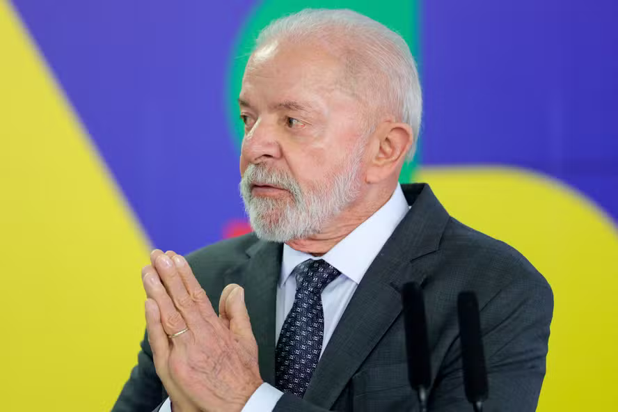 Presidente Lula de terno escuro e gravata, com as mãos juntas, diante de microfones e fundo com as cores da bandeira brasileira.