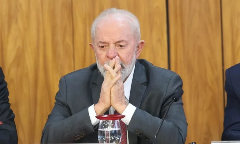 Lula sentado à mesa, de terno escuro e gravata vermelha, com as mãos unidas diante do rosto e expressão séria.