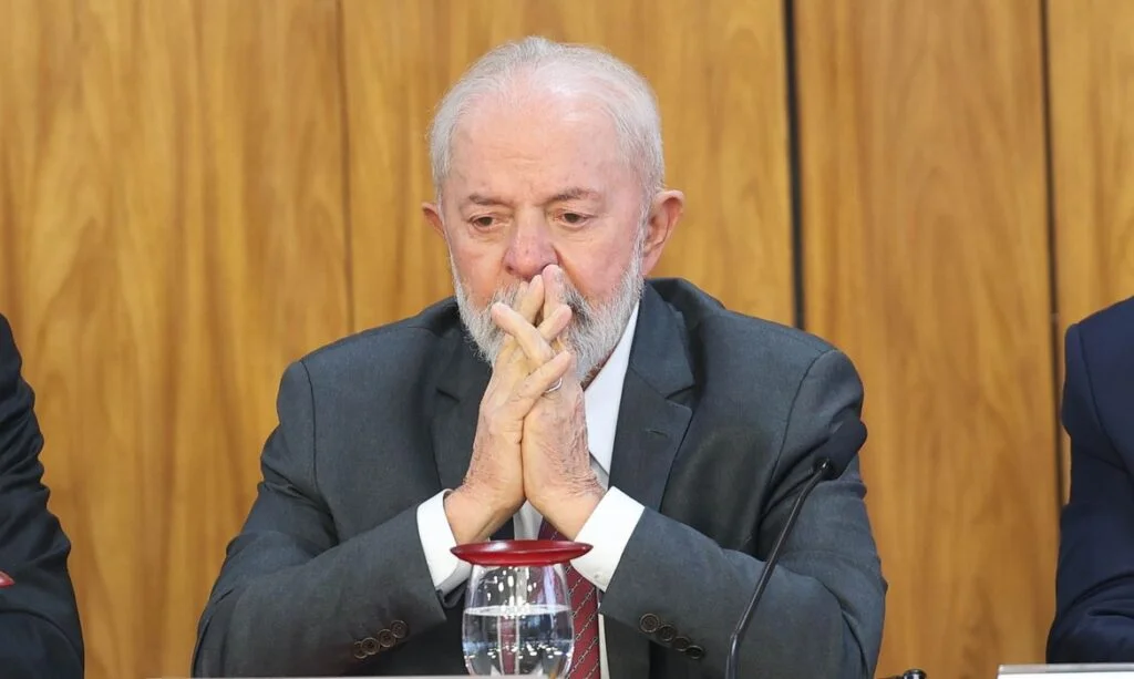 Lula sentado à mesa, de terno escuro e gravata vermelha, com as mãos unidas diante do rosto e expressão séria.