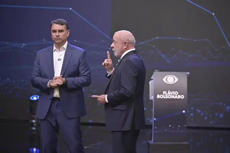 Flávio Bolsonaro, de terno azul e mãos unidas, observa Lula, que gesticula com o dedo indicador levantado durante debate no palco da Band.