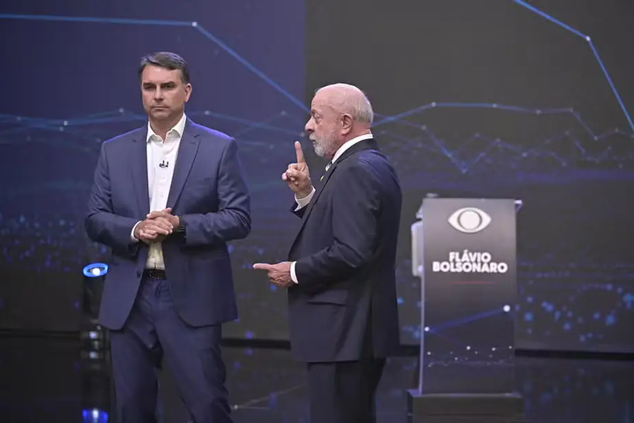 Flávio Bolsonaro, de terno azul e mãos unidas, observa Lula, que gesticula com o dedo indicador levantado durante debate no palco da Band.