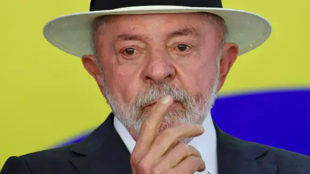 Presidente Lula usando chapéu claro com faixa preta e terno escuro, com o dedo próximo à boca, diante de um fundo amarelo e azul.
