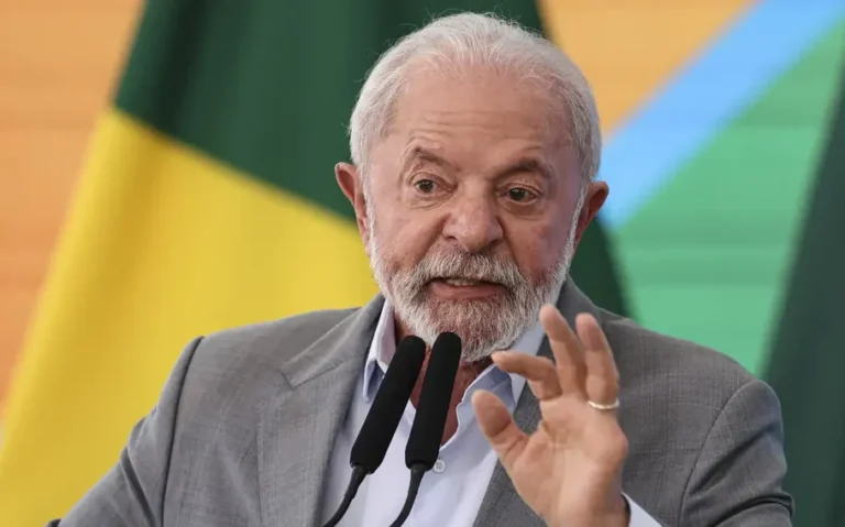 Lula fala diante de dois microfones e gesticula com a mão, com bandeiras brasileiras ao fundo.