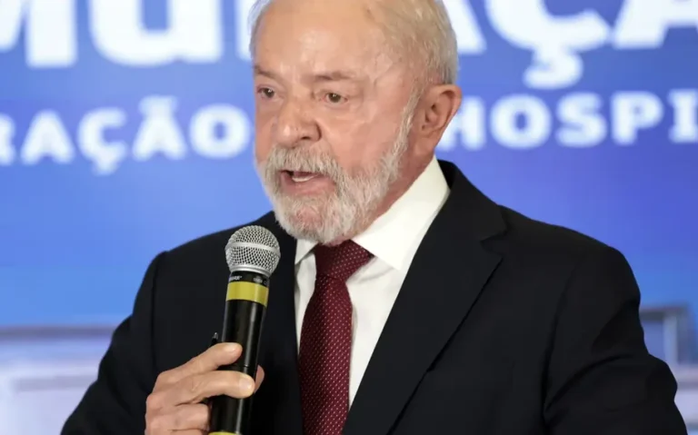 Lula fala ao microfone, de terno escuro e gravata vermelha, diante de painel azul.