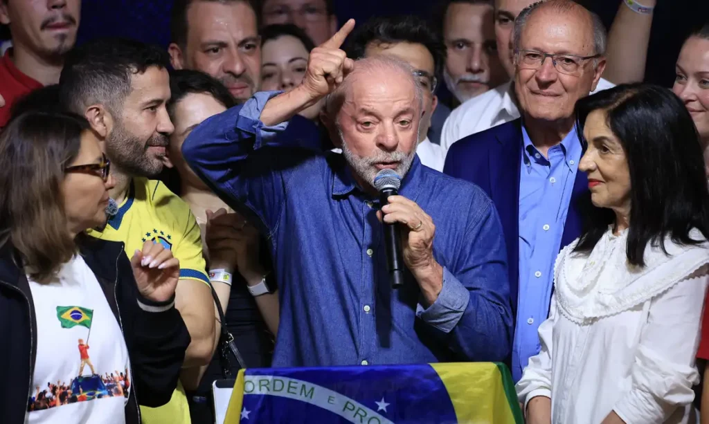 Lula, de camisa azul, fala ao microfone com o dedo indicador erguido, cercado por pessoas e com uma bandeira do Brasil à frente.