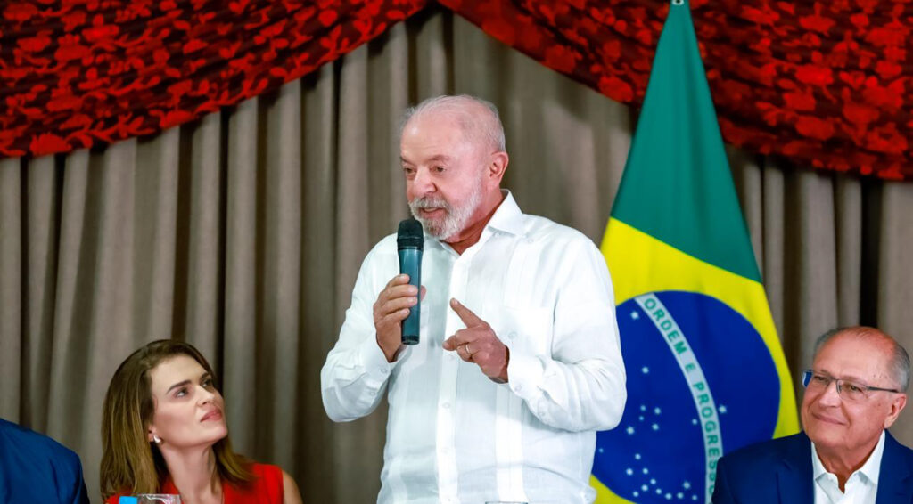 Presidente Lula fala ao microfone durante reunião em Brasília, com a bandeira brasileira ao fundo.