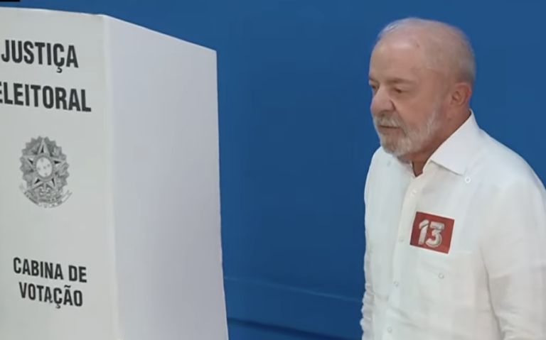 Lula, de camisa branca, ao lado de uma cabine da Justiça Eleitoral, diante de fundo azul.
