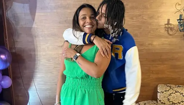 Márcia Gama, de roupa verde, sorri enquanto Oruam, com cabelos longos trançados e jaqueta azul, a abraça e beija seu rosto.