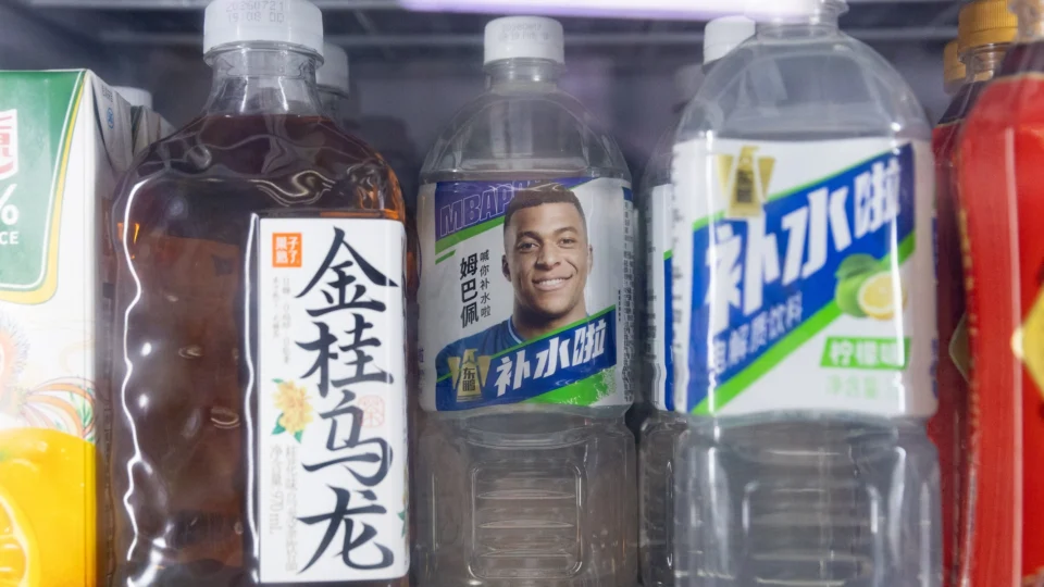 Garrafa de bebida com uma fotografia de Kylian Mbappé no rótulo, posicionada entre outras bebidas com inscrições chinesas.
