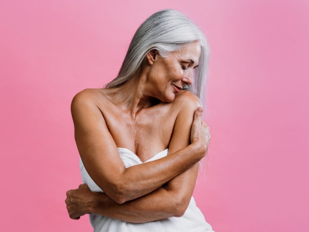 Mulher madura de cabelos longos e grisalhos, envolta em uma toalha branca, abraça o próprio corpo diante de um fundo rosa.