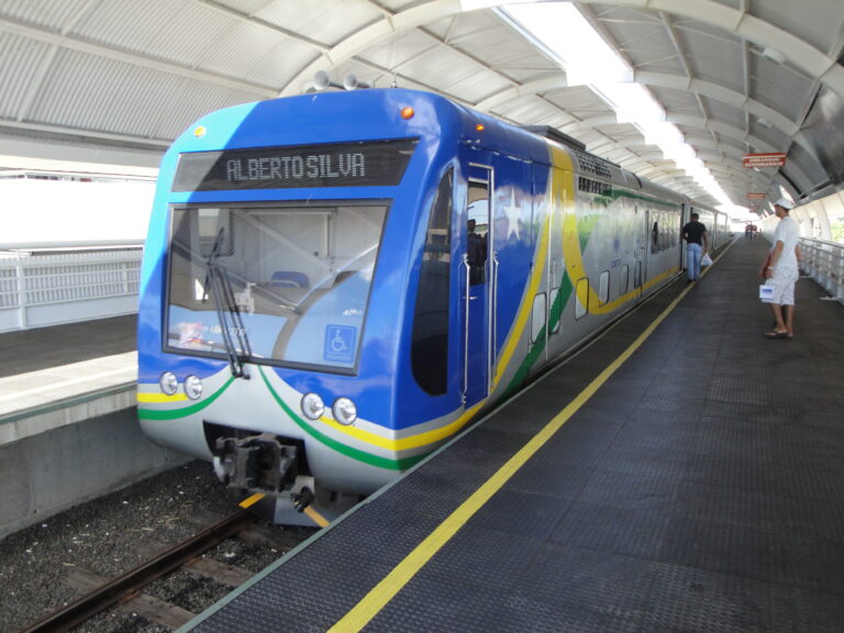 Trem azul, verde e amarelo do Metrô de Teresina parado na plataforma coberta de uma estação, com passageiros ao lado.