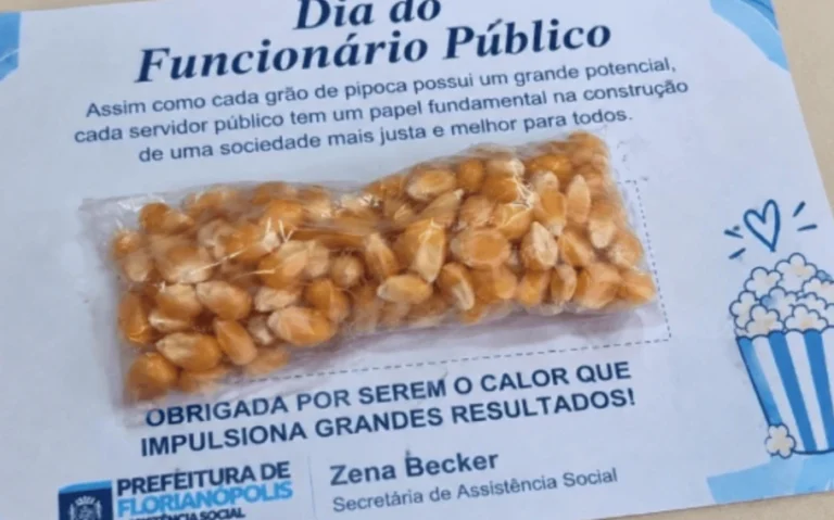 Saquinho transparente com grãos de milho sobre um cartão do Dia do Funcionário Público, com mensagem de homenagem, assinatura de Zena Becker e ilustração de pipoca.
