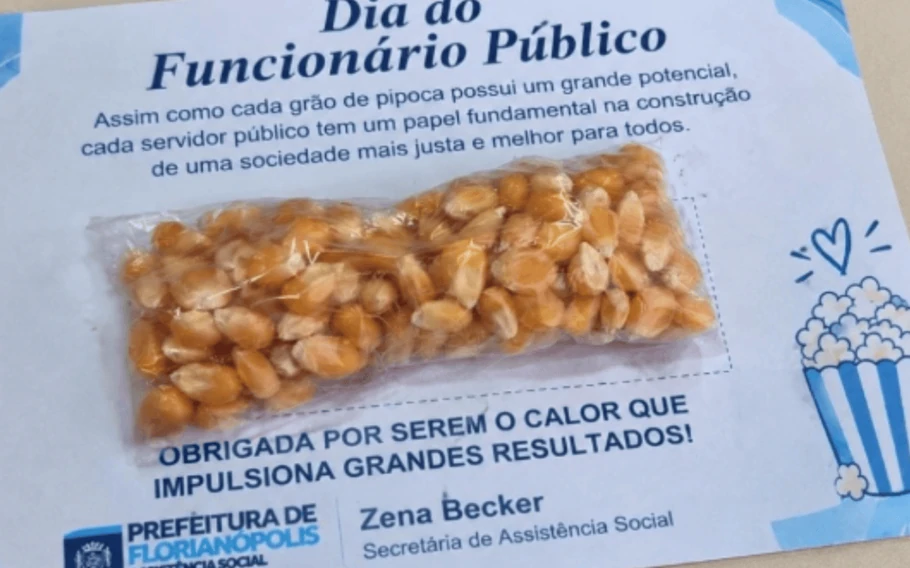Saquinho transparente com grãos de milho sobre um cartão do Dia do Funcionário Público, com mensagem de homenagem, assinatura de Zena Becker e ilustração de pipoca.