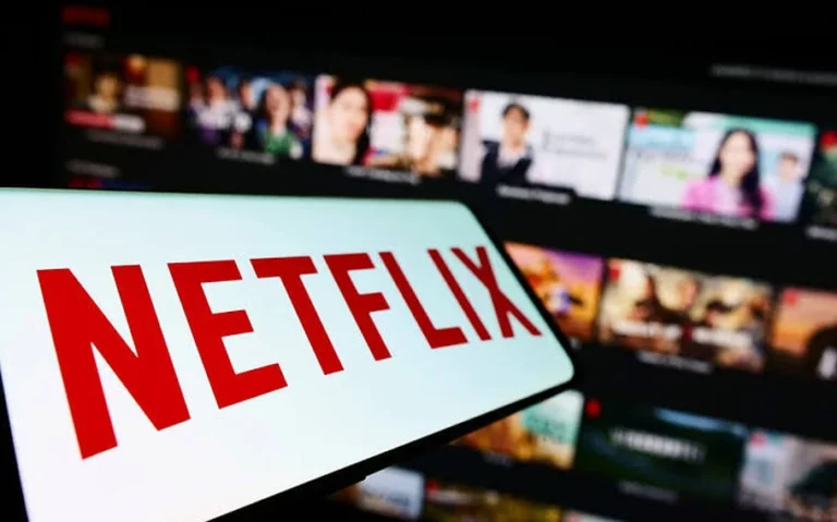 Logotipo vermelho da Netflix sobre fundo branco, com catálogo de filmes e séries desfocado em uma tela ao fundo.