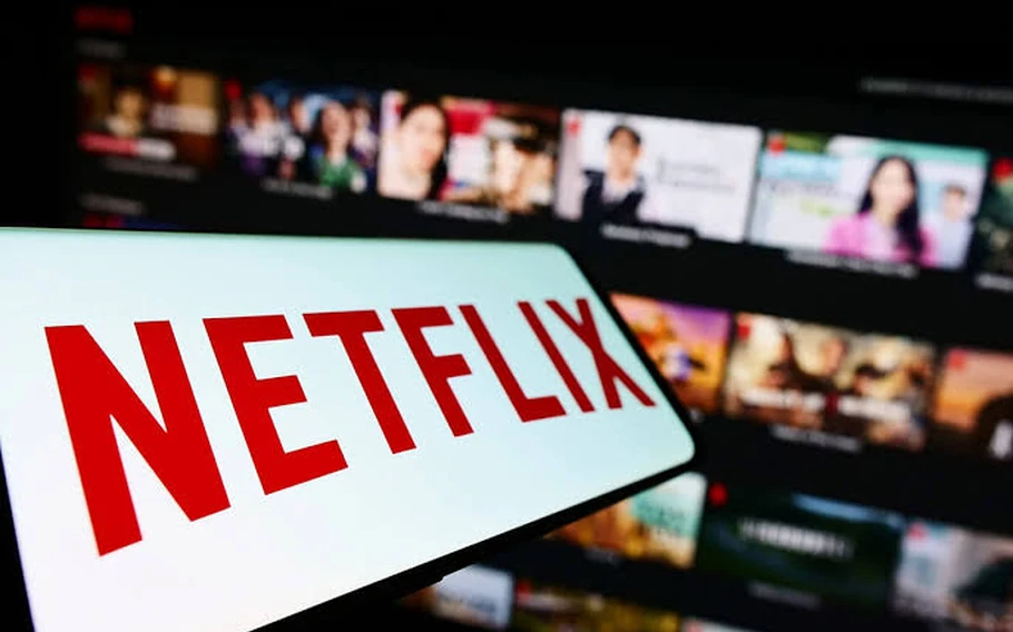 Logotipo vermelho da Netflix sobre fundo branco, com catálogo de filmes e séries desfocado em uma tela ao fundo.
