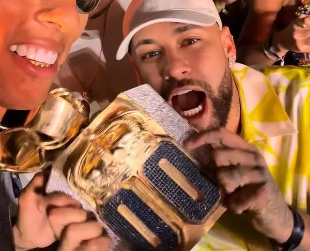 Neymar, de boné claro, e o influenciador Buzeira aparecem sorrindo enquanto exibem um grande colar dourado com pingente em destaque.