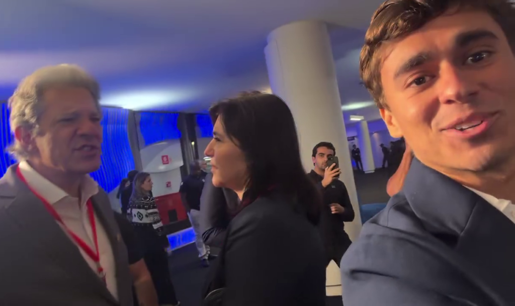 Nikolas Ferreira aparece em primeiro plano, enquanto Fernando Haddad e Simone Tebet conversam ao fundo, nos bastidores do debate da Band.