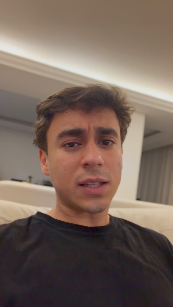 Homem de cabelos castanhos e camiseta preta olha diretamente para a câmera em ambiente interno, com sofá claro e iluminação no teto ao fundo.