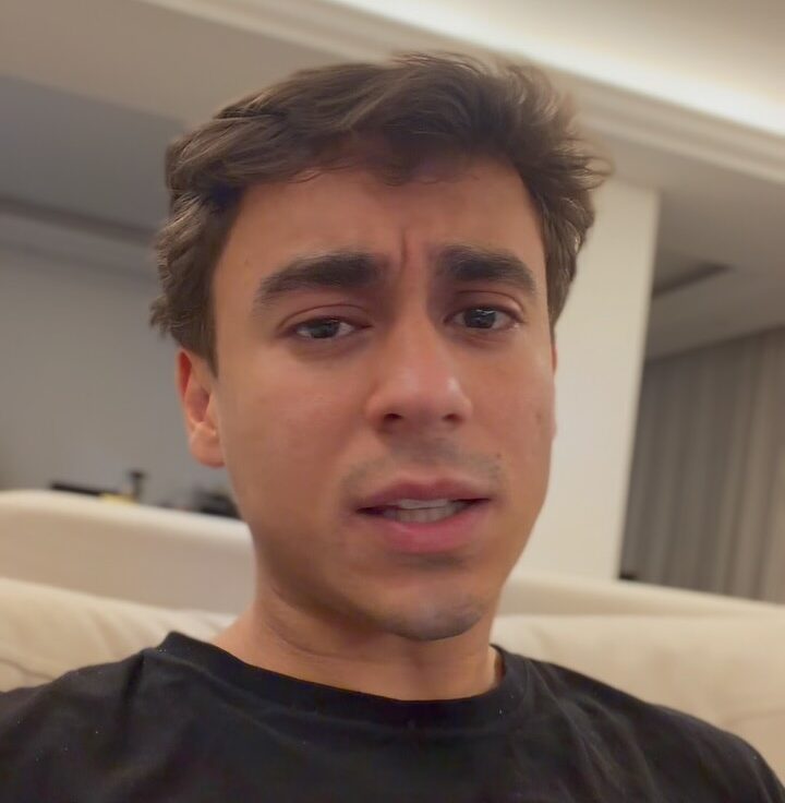Homem de cabelos castanhos e camiseta preta olha diretamente para a câmera em ambiente interno, com sofá claro e iluminação no teto ao fundo.