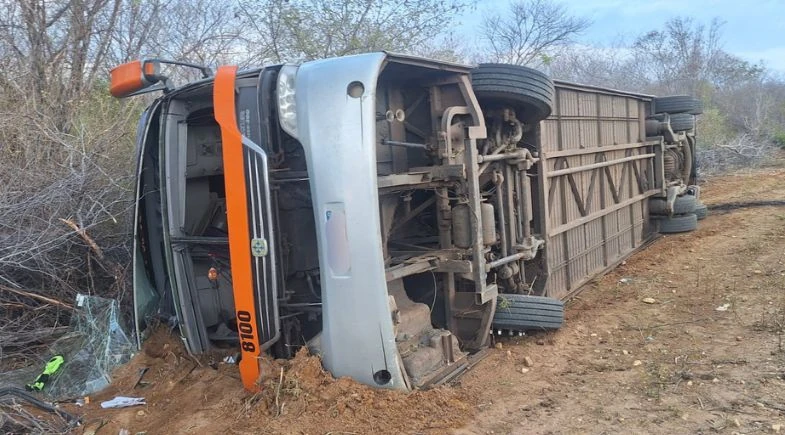 Ônibus cinza com detalhes laranja tombado de lado em terreno de terra, com rodas e parte inferior expostas, próximo à vegetação seca.