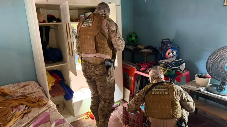 Dois agentes da Força Estadual do Piauí, com uniformes camuflados e coletes táticos, realizam buscas em um quarto com guarda-roupa aberto, cama, mochilas e ventilador.