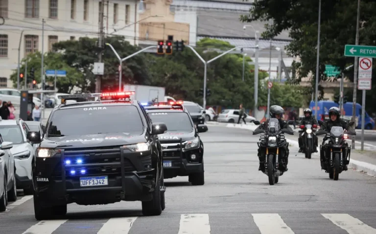 Duas viaturas pretas identificadas como Garra, com luzes de emergência acionadas, e policiais em motocicletas percorrem uma avenida urbana.