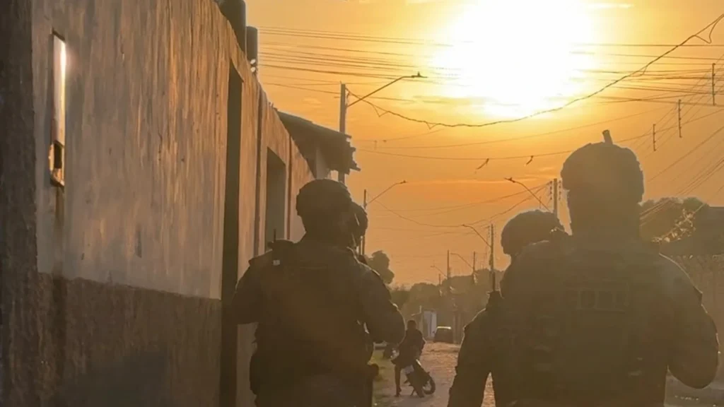Dois policiais com equipamentos táticos caminham de costas por uma rua residencial ao pôr do sol, com casas e fios elétricos ao fundo.