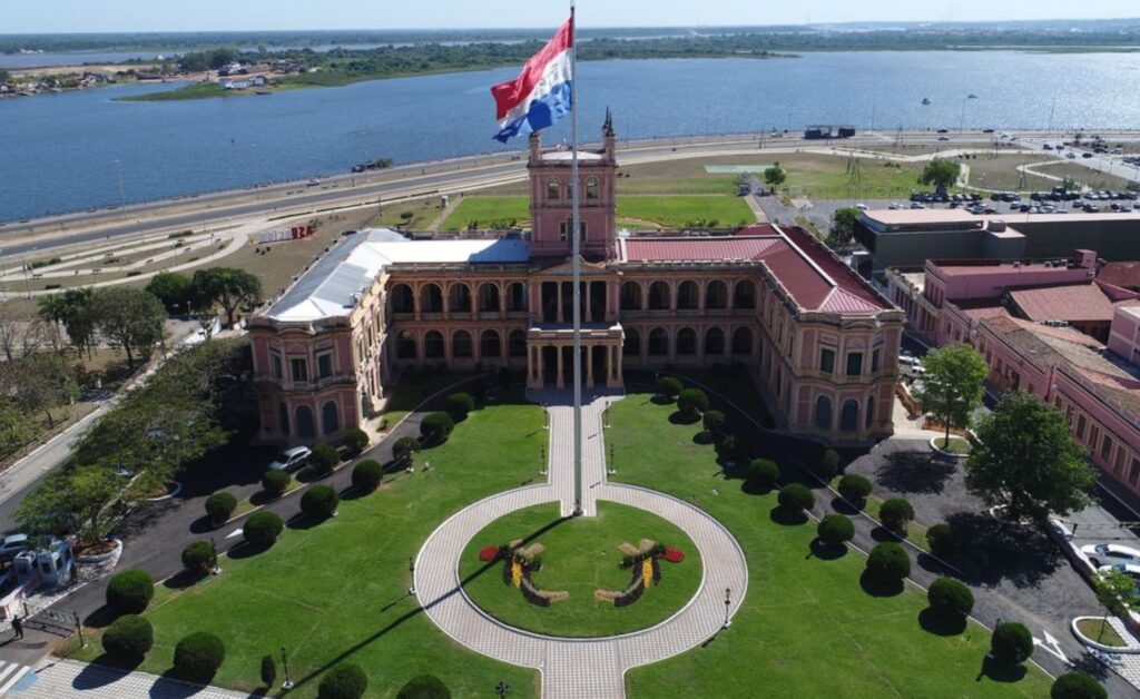 Vista aérea do Palácio de López, em Assunção, no Paraguai, com jardins, bandeira paraguaia e o rio ao fundo.