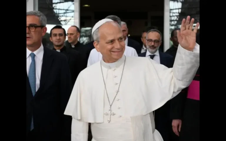 Papa Leão XIV sorri e acena com a mão direita, vestido de branco e acompanhado por religiosos e homens de terno.