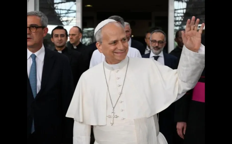 Papa Leão XIV sorri e acena com a mão direita, vestido de branco e acompanhado por religiosos e homens de terno.