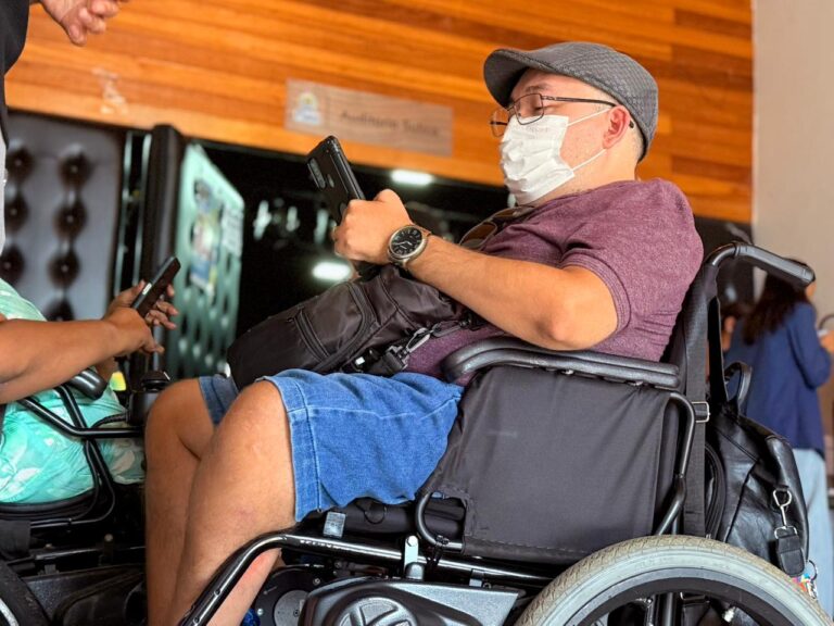Homem em cadeira de rodas, usando boné e máscara, segura um celular em ambiente interno.