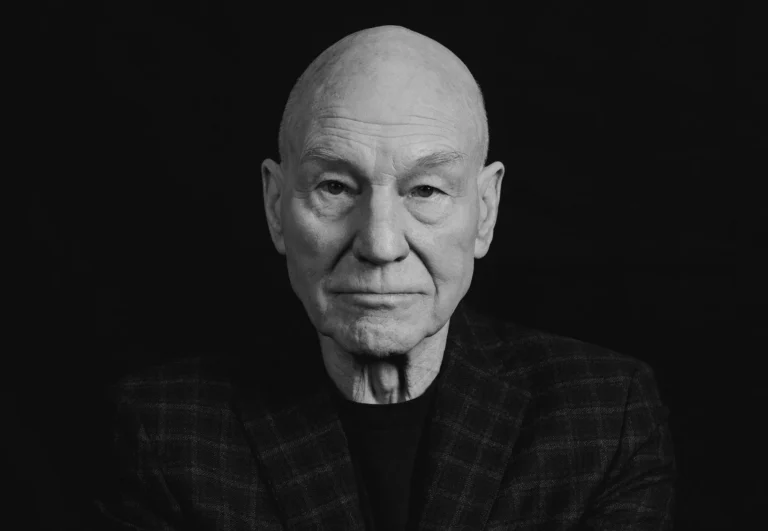Retrato em preto e branco de Patrick Stewart, vestindo blazer escuro diante de um fundo preto.