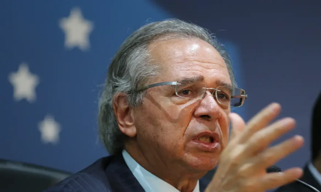 Paulo Guedes, de óculos e terno escuro, fala e gesticula com uma das mãos diante de um fundo azul.