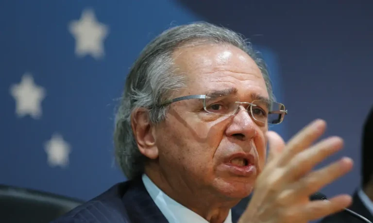 Paulo Guedes, de óculos e terno escuro, fala e gesticula com uma das mãos diante de um fundo azul.