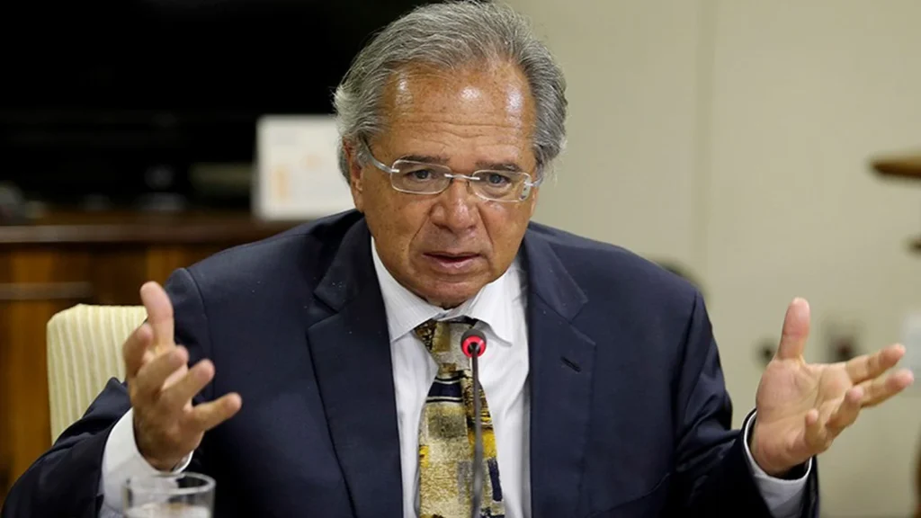 Paulo Guedes, de terno escuro, gravata e óculos, fala ao microfone enquanto gesticula com as duas mãos.