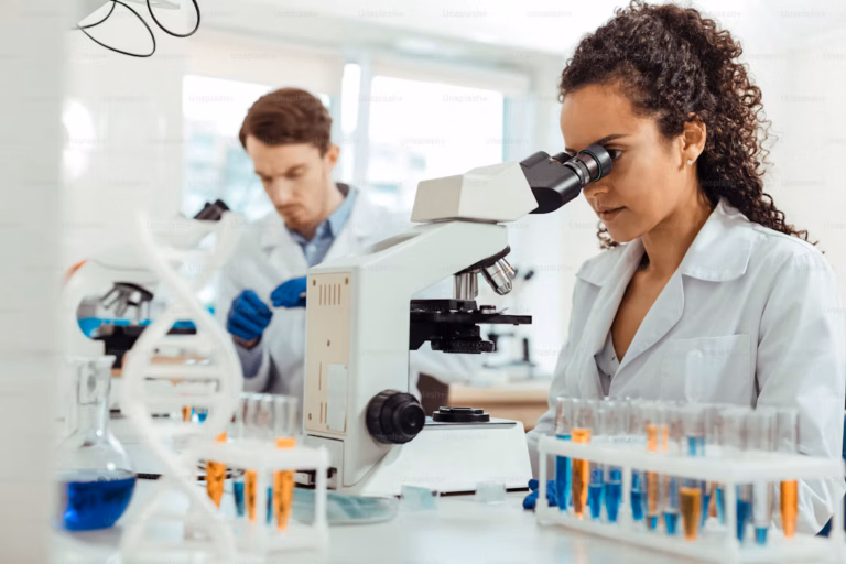 Cientista de jaleco branco observa uma amostra ao microscópio durante atividade de pesquisa em laboratório.
