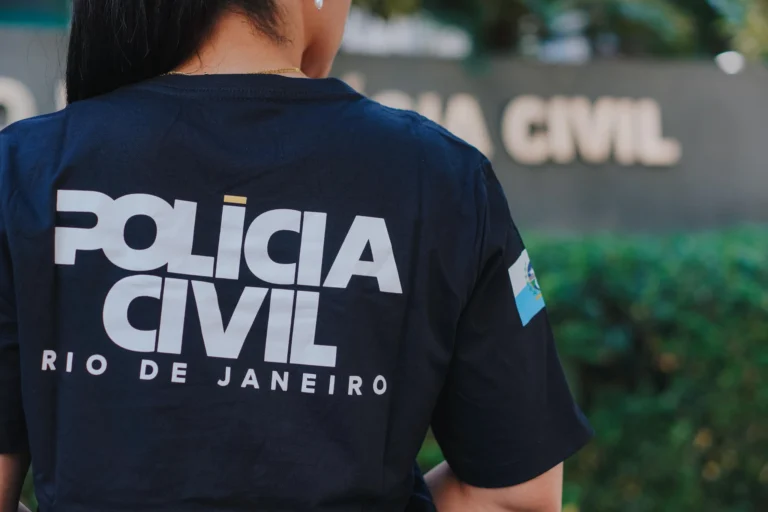 Pessoa vista de costas usa camiseta azul-escura com a inscrição Polícia Civil Rio de Janeiro, diante de uma placa desfocada da instituição.